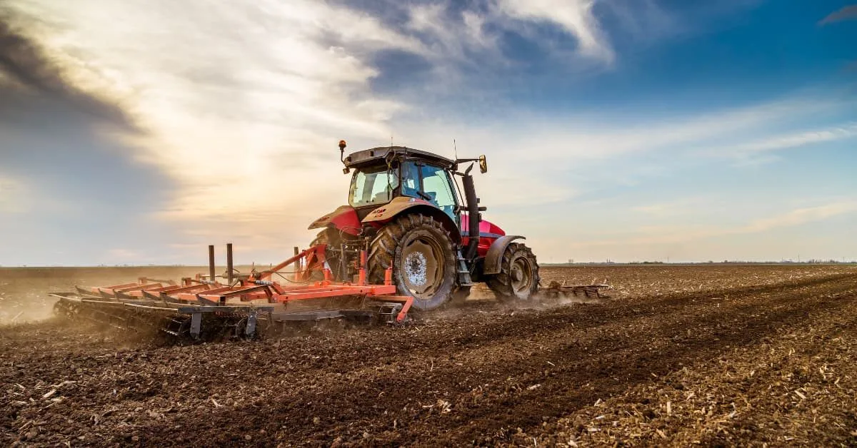 Agricultural tractor working the soil with tractor blade attachment in open field at sunset.